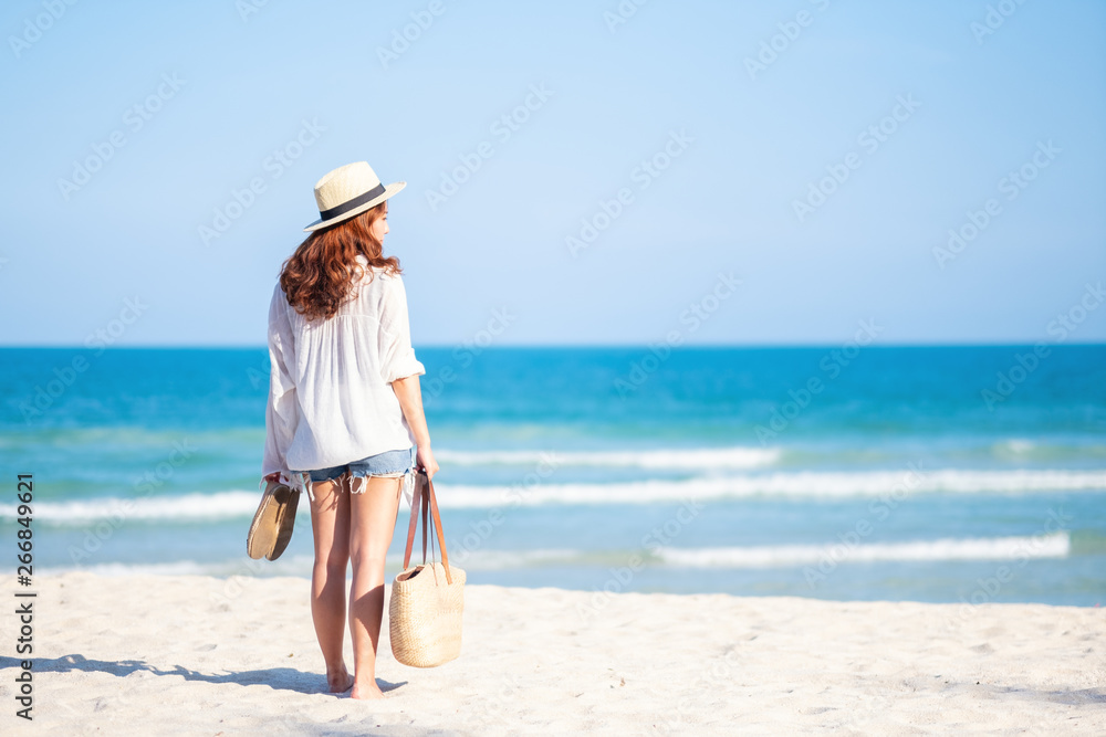 A woman holding bag and shoes while strolling on the beach with the sea and blue sky background