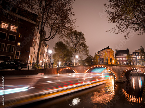Amsterdam, Netherlands. Night view of canals and bridges in Amsterdam, Netherlands. Famous touristic place at night with illuminated houses and long exposure boat ride - Image