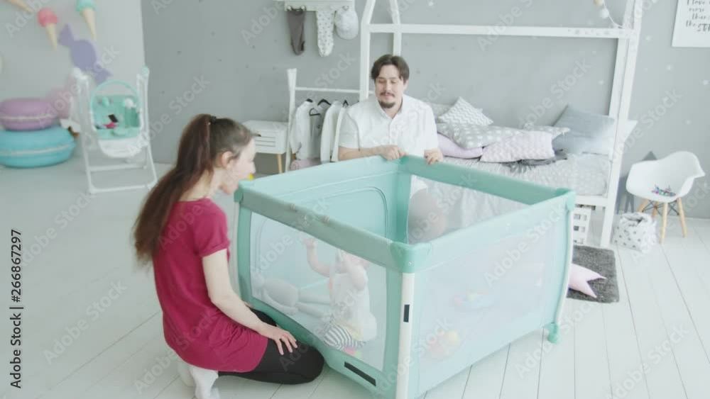 Active little daughter standing by herself pulling on playpen handrails while cheerful mom and dad rejoicing their baby progress. Happy family enjoying domestic leisure and success of cute infant girl Active little daughter standing by herself pulling on playpen handrails while cheerful mom and dad rejoicing their baby progress. Happy family enjoying domestic leisure and success of cute infant girl