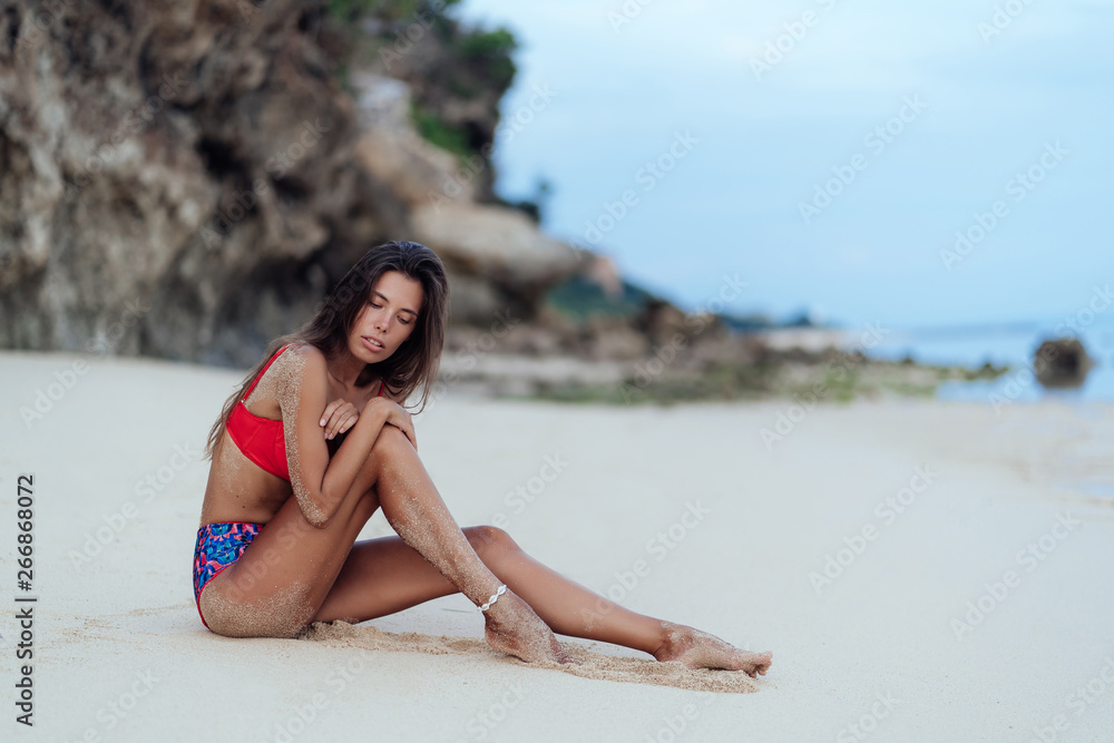 Slender tanned woman in colored swimsuit posing on beach with white sand
