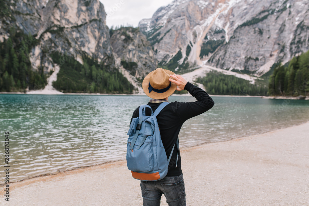 Stylish guy wearing vintage hat decorated with ribbon relaxing on the lake shore and looking at water. Portrait of young man with travel backpack enjoying mountain landscape in cloudy morning.