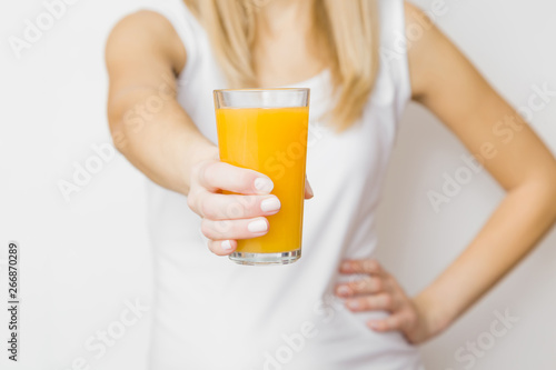 Young blonde woman in white top. Hand holding glass of orange juice on gray background. Tasty, healthy drink. Front view. Close up.