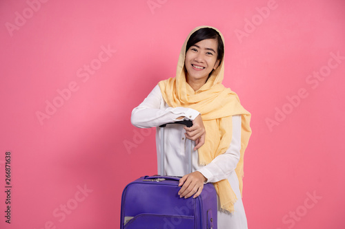 smiling attractive asian muslim woman with scarf looking at camera and holding suitcase