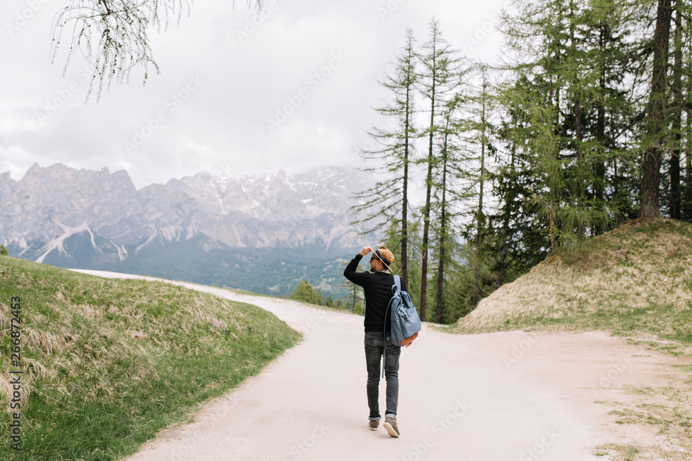 Young man in black shirt carrying backpack spending time outdoor enjoying nature views of Italy in weekend. Full-length portrait from back of traveling guy in hat walking down the street to mountain.