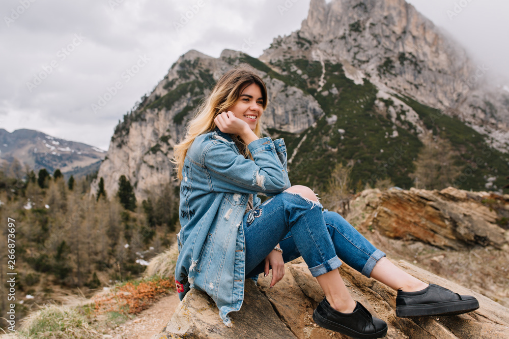 Naklejka premium Dreamy fair-haired girl wearing denim attire resting on the rock after hard climbing and posing touching her face. Attractive stylish young woman with charming smile sitting on stone on mountain view.
