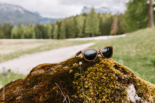 Photo of green landscape with mountains and forest in distant with trendy sunglasses in foreground. Blur picture of nature view with forgotten accessory on stone overgrown with moss.