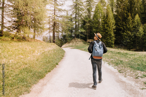 Male traveler with big blue backpack going to forest thicket and looks around with interest. Young man in elegant hat enjoying nature views during trip to pinery in sunny warm day.