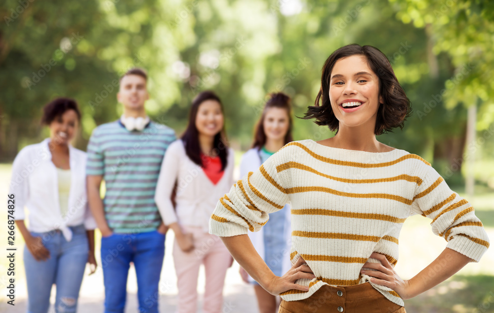 Obraz premium people concept - happy smiling young woman in striped pullover with hands on hips over group of friends in summer park background
