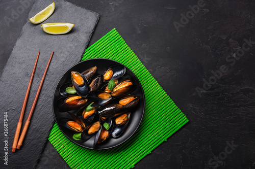 Seafood mussels, basil leaves in a black plate with lemon and chopsticks on green bamboo mat and stone table