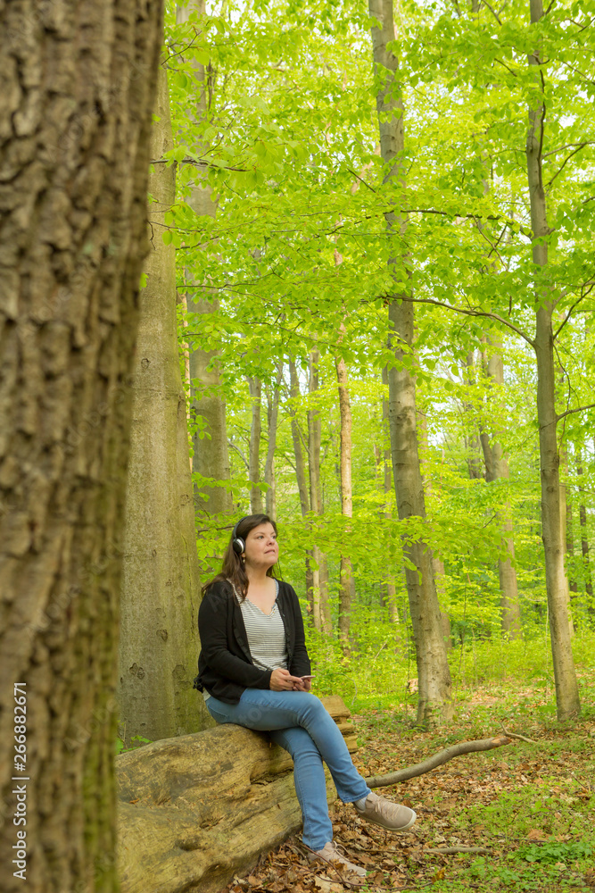 Woman sits on la log of a public park to listen to music with headphones Woman sits on la log of a public park to listen to music with headphones