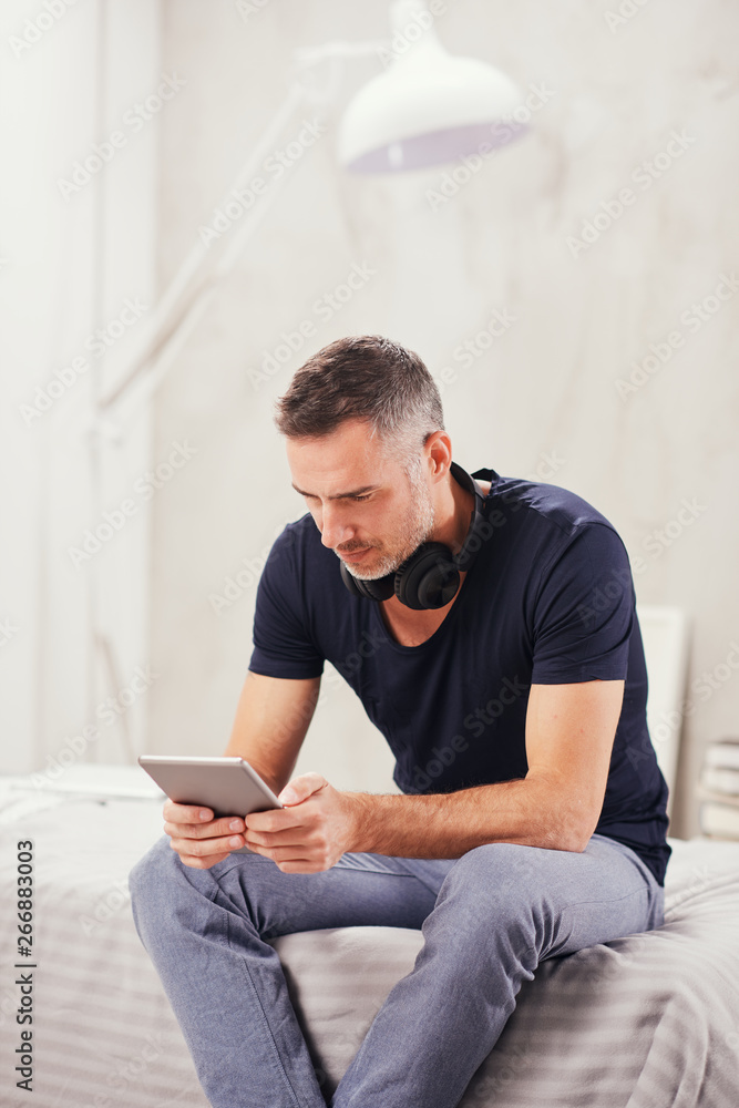 Caucasian man in forties sitting on bed in bedroom and using tablet.