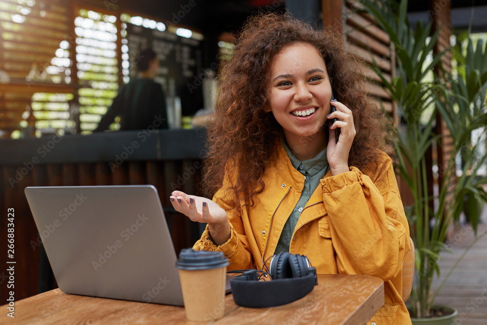 Photo of young dark skinned curly woman siting on a cafe terrace, wearing in yellow coat, works at a laptop, drinking coffee, smiling and talking on the phone with a friend. - obrazy, fototapety, plakaty 