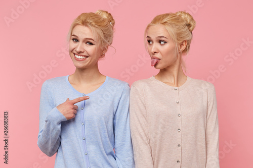 One of beautiful young blonde twins is fooling around and showing her tongue, her sister smiles broadly and pointing at her isolated over pink background.