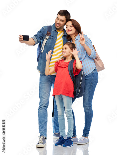 family, tourism and travel concept - happy smiling mother, father and little daughter with backpacks taking selfie by smartphone over white background