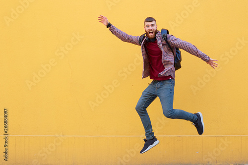 Happy smiling young attractive ginger bearded man jumping, wearing in basic clothes with backpack. Looking at the camera with wide open mouth over a yellow wall with copy space.