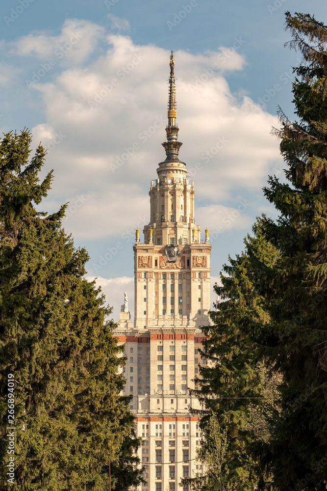 The building of the Moscow State University is surrounded by green coniferous trees against the blue sky with clouds