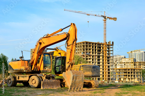 Heavy tracked excavator at a construction site on a background of a residential building and construction cranes on a sunny day against the backdrop of a sunset and blue sky with clouds