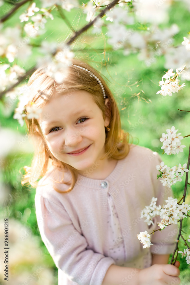 Portrait of cute and pretty little girl in spring blooming trees. Adorable little girl smiling in blooming garden on sunny day. Child and flowers. Childhood & natural tenderness. Children's day.