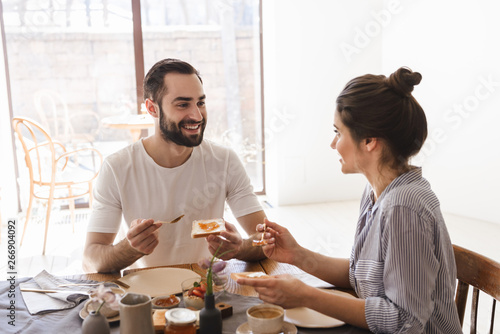Image of satisfied brunette couple eating breakfast together while sitting at table in apartment