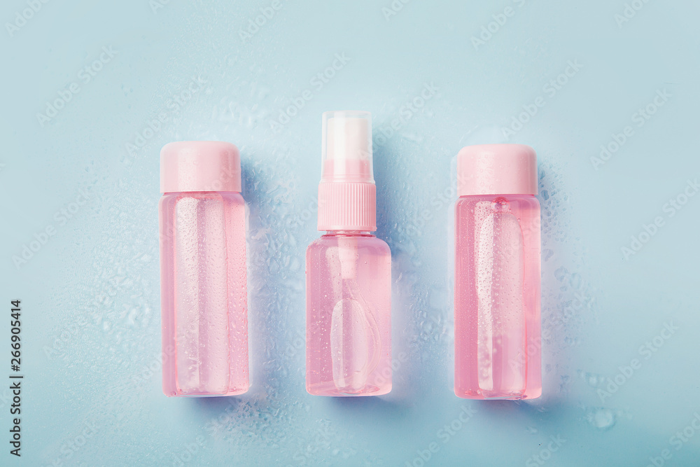 Three bottles of pink tonic or lotion sprinkled with water on blue background. Freshness and body care. Female cosmetics. Micellar water. Travel set of face care cosmetic Three bottles of pink tonic or lotion sprinkled with water on blue background. Freshness and body care. Female cosmetics. Micellar water. Travel set of face care cosmetic