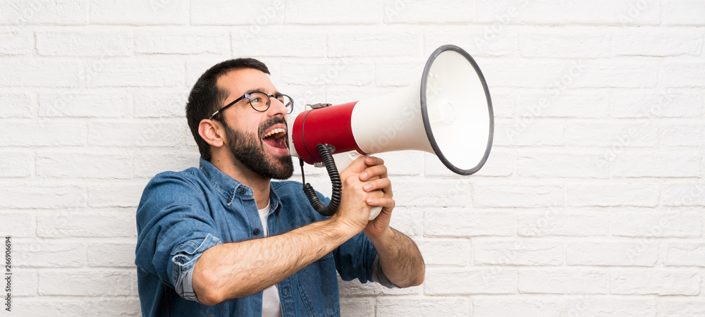 Handsome man with beard over white brick wall shouting through a megaphone - obrazy, fototapety, plakaty 
