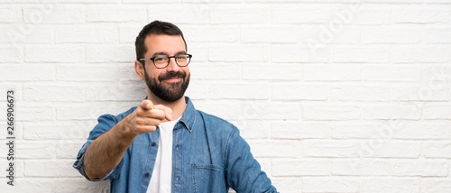 Handsome man with beard over white brick wall points finger at you with a confident expression