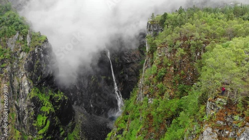 Tourist woman standing on cliff at Voringsfossen waterfall, waving to camera. Mabodalen canyon Norway. National tourist Hardangervidda route,  Eidfjord sightseeing tour photo