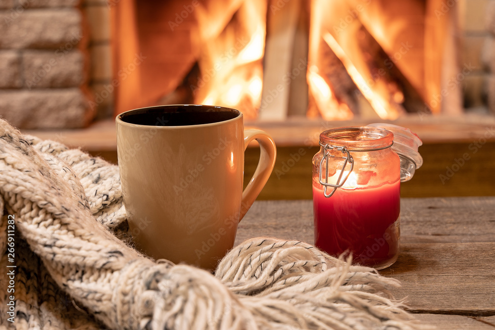 Big mug with hot tea, and a candle , wool scarf, near cozy fireplace, hygge, home sweet home. - obrazy, fototapety, plakaty 
