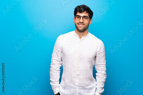 Young man over isolated blue wall with glasses and happy