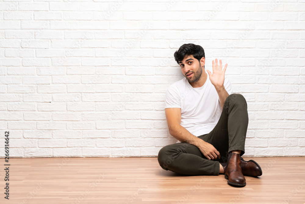 Young man sitting on the floor saluting with hand with happy expression Young man sitting on the floor saluting with hand with happy expression
