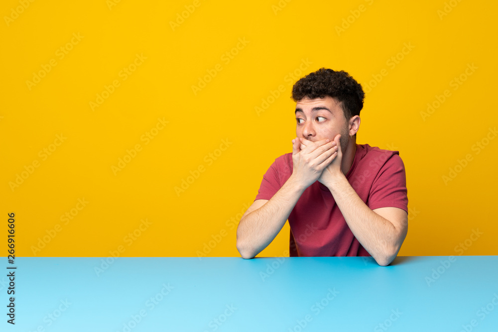 Young man with colorful wall and table covering mouth and looking to the side Young man with colorful wall and table covering mouth and looking to the side