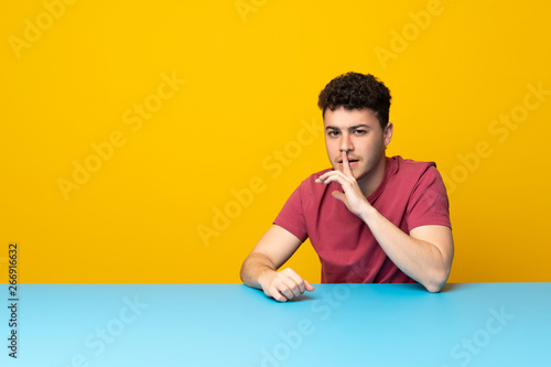 Young man with colorful wall and table showing a sign of silence gesture putting finger in mouth