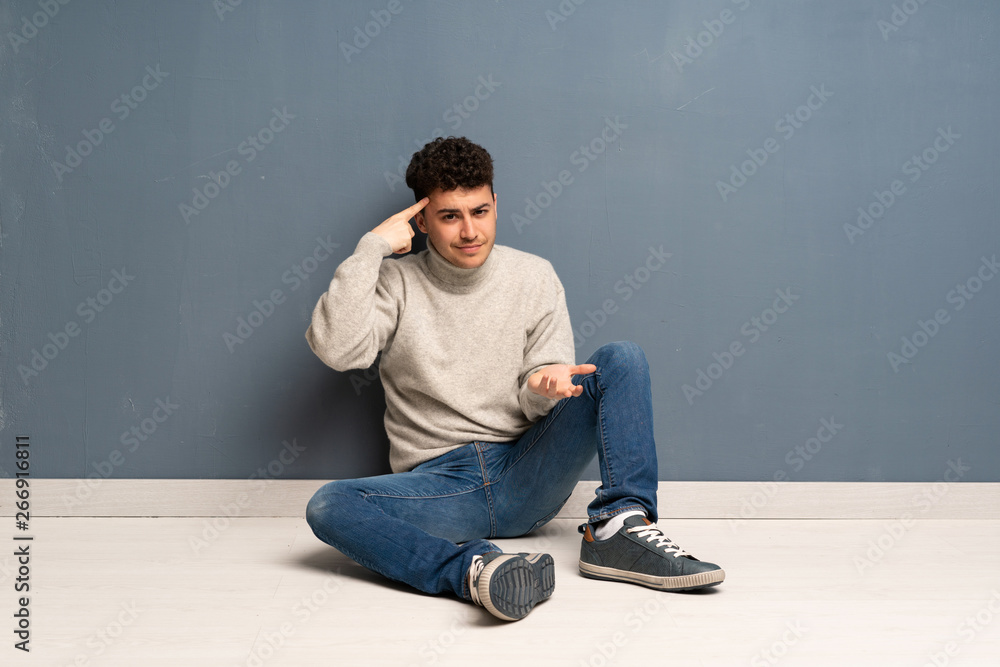 Young man sitting on the floor making the gesture of madness putting finger on the head Young man sitting on the floor making the gesture of madness putting finger on the head