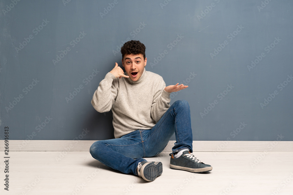 Young man sitting on the floor making phone gesture and doubting Young man sitting on the floor making phone gesture and doubting