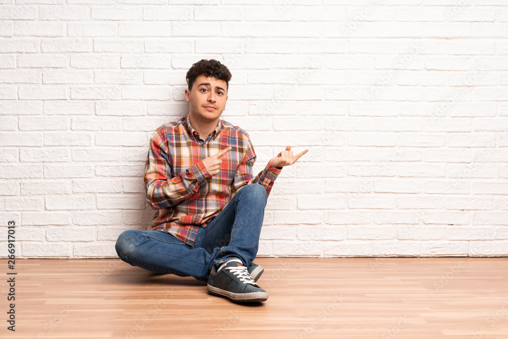 Young man sitting on the floor frightened and pointing to the side Young man sitting on the floor frightened and pointing to the side