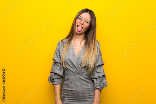 Young woman with glasses over yellow wall showing tongue at the camera having funny look