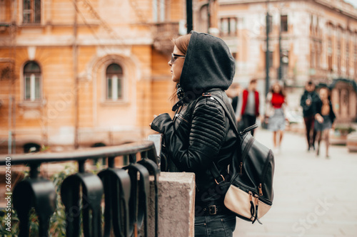 Young woman wearing jeans, a leather jacket with hood and small backpack – Casually dressed millennial exploring and walking through the city – Concept image for travel or tourism