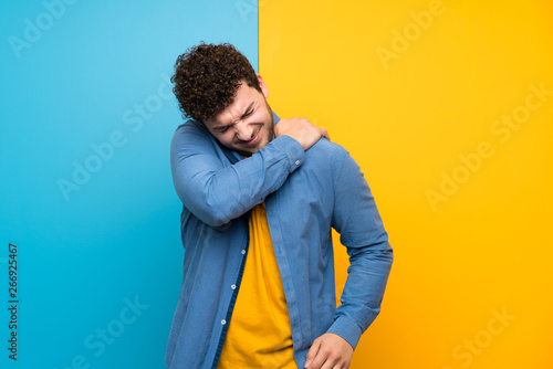 Man with curly hair over colorful wall suffering from pain in shoulder for having made an effort © luismolinero