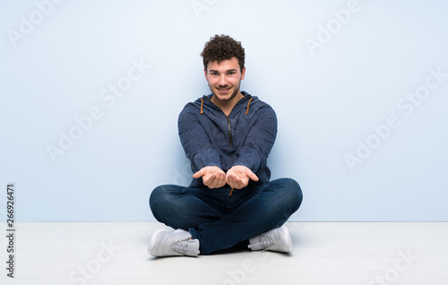 Young man sitting on the floor holding copyspace imaginary on the palm to insert an ad © luismolinero