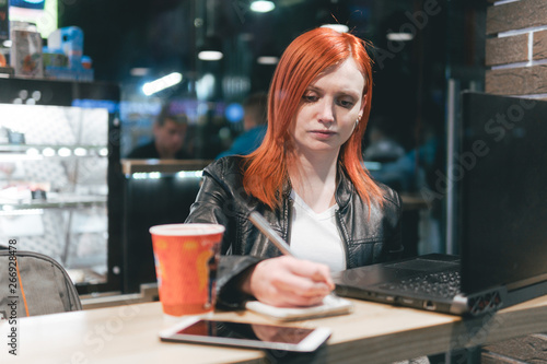 Businesswoman  girl holding a pen  writing in a notebook  laptop in cafe  smartphone  pen  use computer. Freelancer works remotely. Online marketing  education for adulte. Night city  dark theme. tea