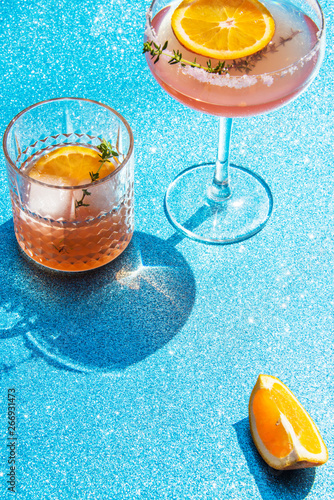Grapefruit and thyme cocktails, refreshing drinks with ice on a light blue background. Summer garden party still life with orange and cold drinks, trendy retro style with artistic shadows.