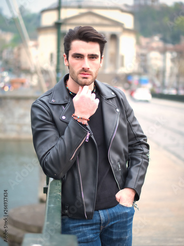 One handsome young man in urban setting in modern city, standing, wearing black leather jacket and jeans, looking at camera