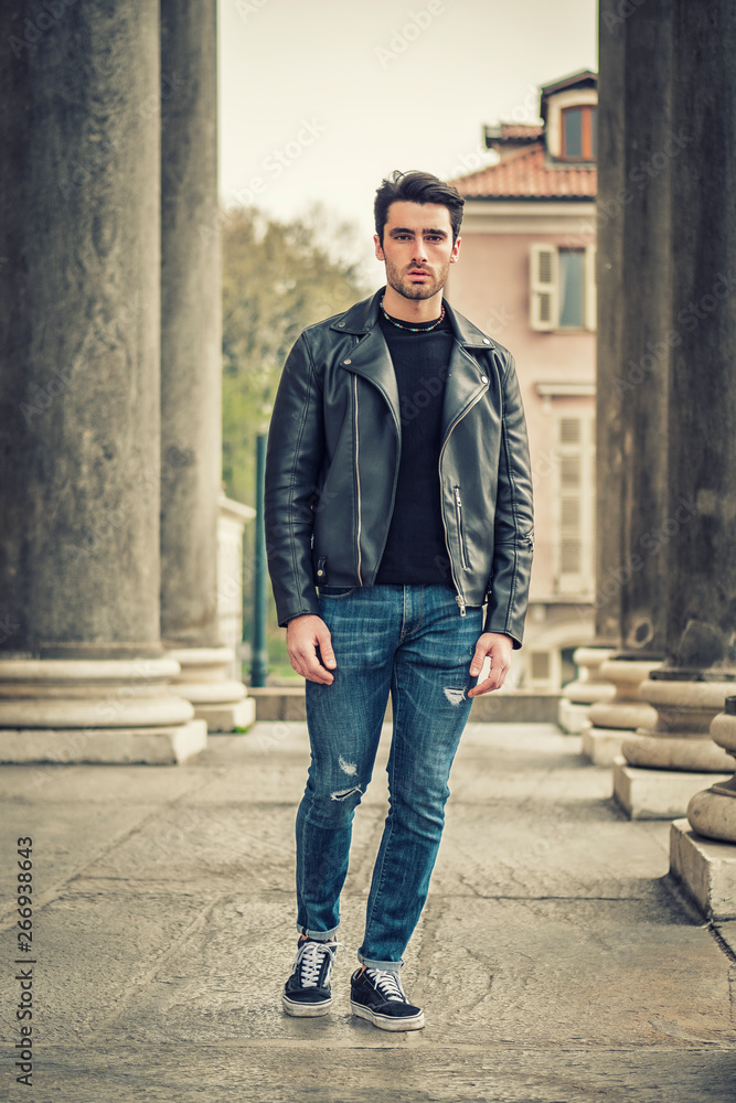 One handsome young man in urban setting in modern city, standing, wearing black leather jacket and jeans, looking at camera One handsome young man in urban setting in modern city, standing, wearing black leather jacket and jeans, looking at camera