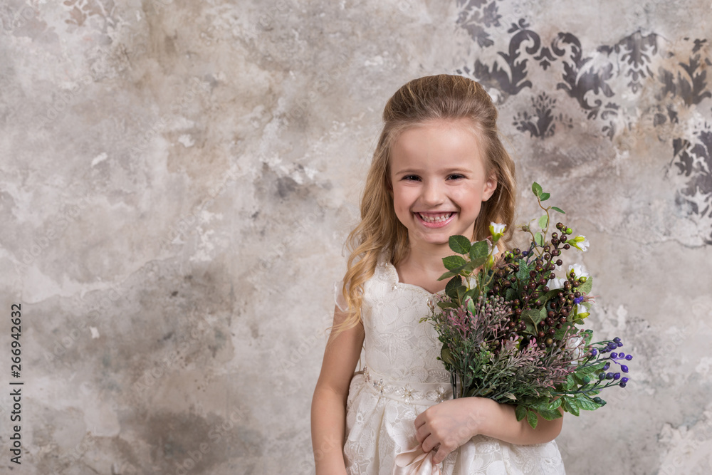 Portrait of a little attractive girl in a white dress with a bouquet in her hands against the background of a grunge wall.