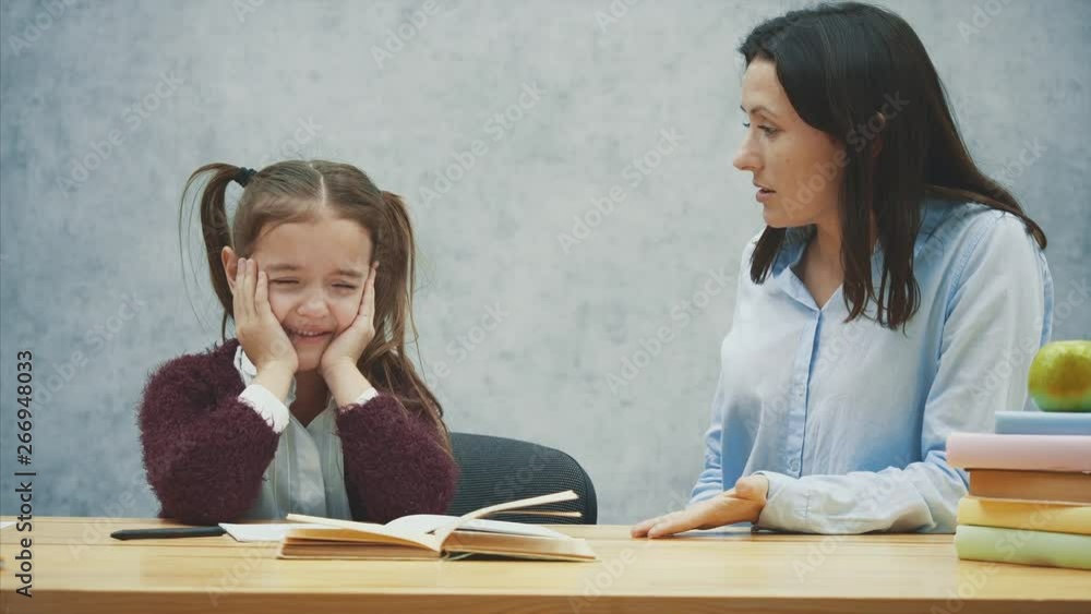 A girl on a gray background sits at the table and cries. During this, my mother is quarreling with her daughter. Concept of relations. A girl on a gray background sits at the table and cries. During this, my mother is quarreling with her daughter. Concept of relations.