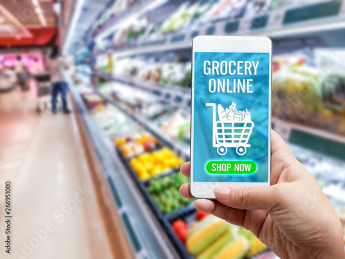 Online grocery shopping concept: Woman hand holding smart phone for ordering food onscreen with icon media on supermarket background. Business and technology for lifestyle in city.