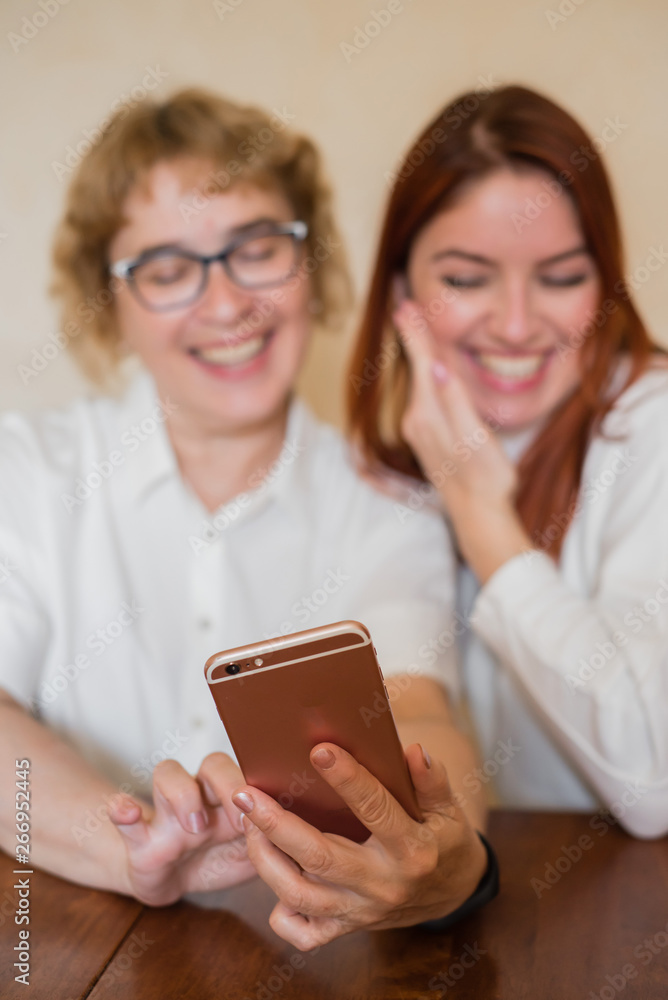 An elderly mother and daughter dressed in light clothes are smiling and taking selfies. Mom and daughter are photographed on the front camera of the smartphone while sitting at home at the table. An elderly mother and daughter dressed in light clothes are smiling and taking selfies. Mom and daughter are photographed on the front camera of the smartphone while sitting at home at the table.