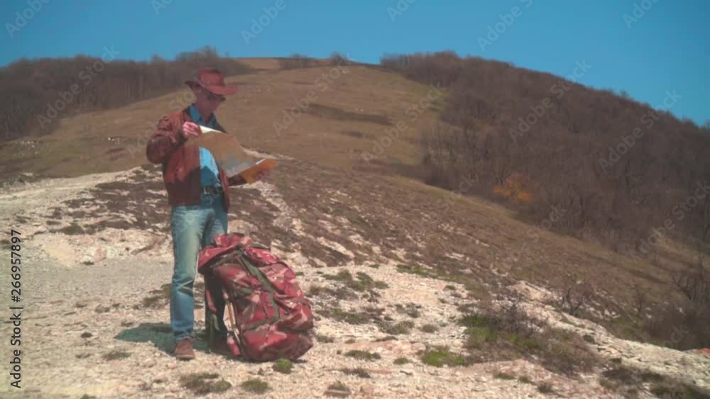 A man in a cowboy hat, leather jacket, blue jeans and glasses is standing in the mountains and considering a traveler map. Background of mountains and sky. A man in a cowboy hat, leather jacket, blue jeans and glasses is standing in the mountains and considering a traveler map. Background of mountains and sky.