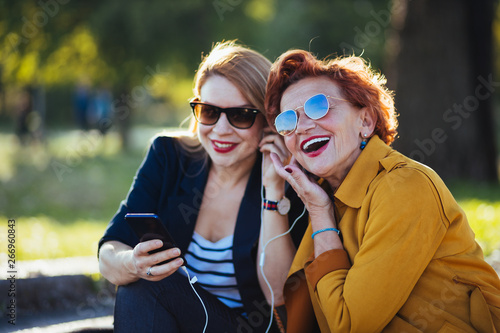 Mature mother and adult daughter listening to the music on smartphone in the park