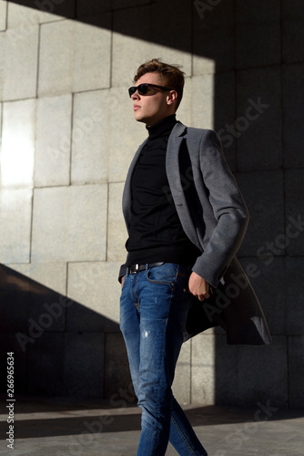 Fashionable young adult man in sunglasses. Posing in grey elegant jacket, black polo neck, jeans trousers and brown mocassin shoes. Fashion trends.